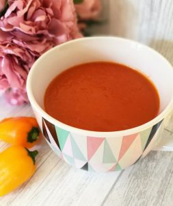 Spicy tomato pepper & chorizo soup in a bowl with 2 small yellow peppers next to it.