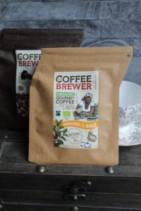 Growers Cup Review - A light brown coffee bag sitting on a dark brown chest with a dark brown coffee bag and a white teacup behind it. The label on the front coffee bag says it's Organic Gormet Coffee from Ethiopia