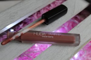 January Makeup Favourites -A brown tube of lipstick with Huda Beauty logo on it. Tthe applicator is resting on top. Both are sat on grey slatted wood with pink showing through.