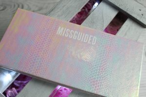 January Makeup Favourites - A rectangle package resting on a grey slatted wood background.The rectangle palette is a pink/blue holographic snakeskin with MISSGUIDED written along the top edge.