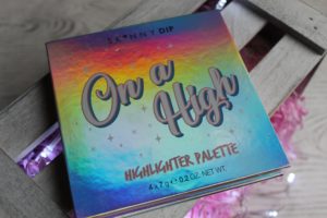 January Makeup Favourites - A square, rainbow coloured box with On A High Highlighter Palette written on it. IT is sat on a grey, slatted wood background with a little pink showing through.