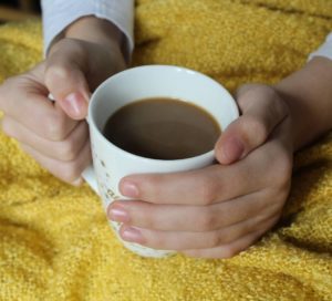 Growers Cup Review - A white mug filled with coffee, hands holding the handle and wrapped around the mug. The mug is sat in someones lap that is covered with a mustard coloured blanket.