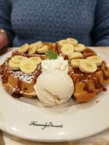 Heavenly Desserts Colchester - A white plate with the company name in black then a large round waffle covering the rest of the plate. A ball of white ice cream in front and chopped banana on top with toffee sauce drizzled over it