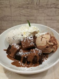 Heavenly Desserts Colchester - A white bowl containing chocolate brownie chunks with chocolate sauce drizzled over them, a white ice cream ball, a brown one and some whipped cream topped with a small mint leaf 