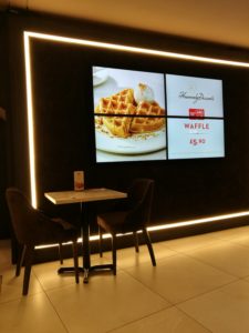 Heavenly Desserts Colchester - A black wall with a white light framing the edge and 4 TV screens in the middle with one big picture of waffles showing on them and a small table and 2 chairs to the left