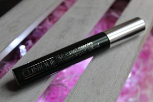 January Makeup Favourites - a black mascara tube with a silver lid. Written in silver along the tube is "Clinique high impact mascara." The mascara is laying across grey, wooden slats with pink showing inbetween.