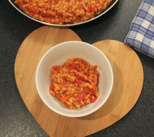 Roasted Sweet Red Pepper And Tomato Risotto - a white bowl containing orange and red risotto, sat on a heart shaped wooden board. A pan of risotto sits to the top of the picture and a blue and white gingham tea towel is to the right.