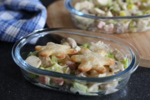 Chicken, bacon and leek pot pie - a glass pie dish in the foreground containing chunks of pie filling with 2 flower shaped bits of pastry sat on top. In the background is a blue and white teatowel folded up and another glass pie dish sat on a wooden chopping board.