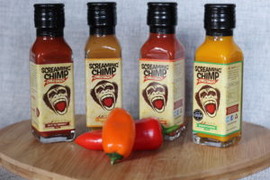 Screaming Chimp Chilli Sauce - 4 rectangular bottles in a row, stood on a wooden board with 2 small peppers in front of them.