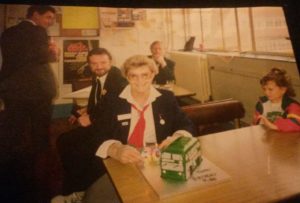 Loneliness in the elderly - An old photo of a lady sat at a table with a cake in the shape of a bus in front of her, a young child to her left and some men looking on from behind 