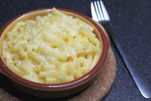 Cheats Mac and Cheese - a bowl of cheese topped pasta on a cork mat and a fork lying next to it on a black worktop.