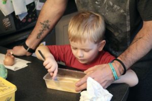 Things to do with the kids in January - 4yo boy with a fork in hand stirring up his scrambled eggs clearly being helped by his Dad who's arms are either side of the boy.