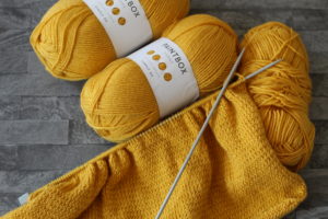 5 New Things To Learn - Two and a bit mustard yellow balls of yarn with knitting needles resting on top with some knitting already done on the bottom needle.