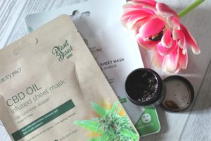 Form New Skincare Habits - 2 large packets of sheet masks lying on a white wood background. An open black pot on top of them that's open and contains a brown sticky looking stuff. The pink flower is by the side of all of these.