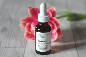 Form New Skincare Habits - A small brown glass bottle with a white label and white dropper top standing in front of a pink flower on a white wood background.