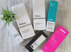 6 boxes of DECIEM products layed out on a white wood background with a small spiky plant to the left.