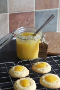 Gluten free, vegan thumbprint biscuits - a jar of bright yellow lemon curd and in front is 4 biscuits sat on a cooling rack