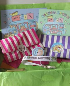 Inside the Sugar & Sloth Tiny Party Club box. 2 colourful postcards, a pink striped bag, a purple stripped bag, a badge and a bracelet