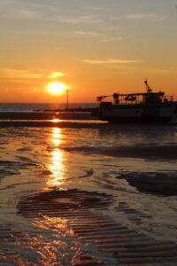 Sea Safety - a sunrise picture with a reflection of the sun in the sand and a silhouette of a boat beached on the sand.