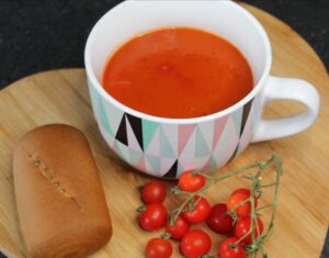 simple tomato and basil soup in a large cup/bowl on a chopping board with small tomatoes on the vine in front and a bread roll to the side