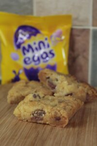 Mini Egg Cookie Bar cut up and sitting stacked on a wooden board. A pack of Mini Eggs can be seen in the background